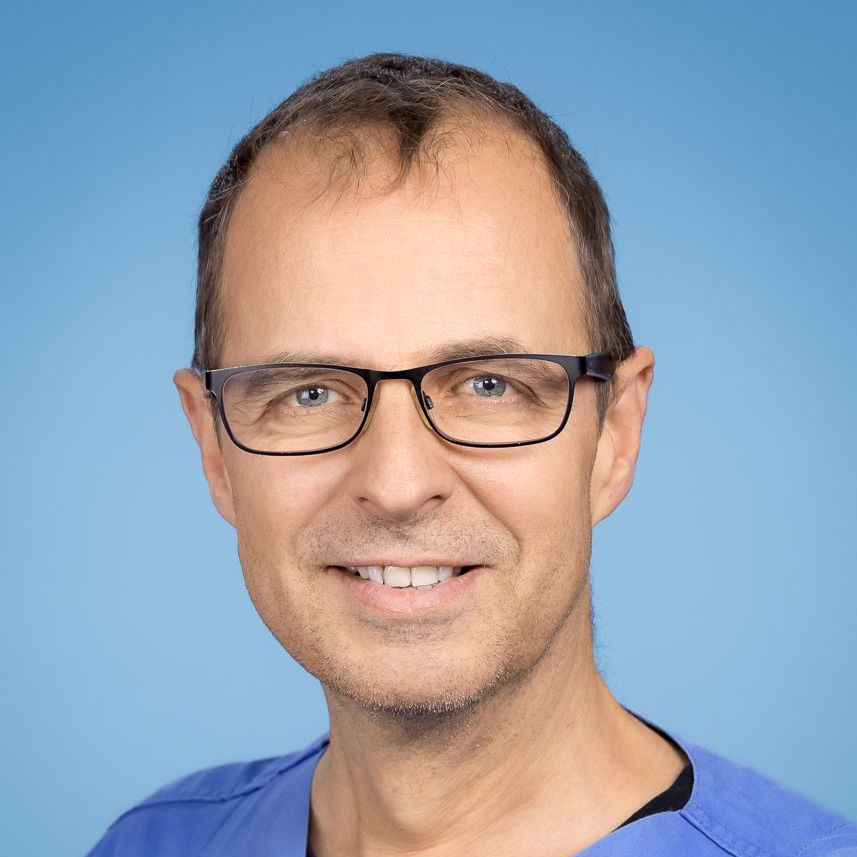 Ein Mann mit Brille und blauem OP-Kittel lächelt in die Kamera.