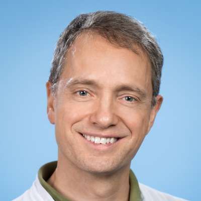 Ein Mann in einem weißen Kittel und einem grünen Hemd lächelt in die Kamera.