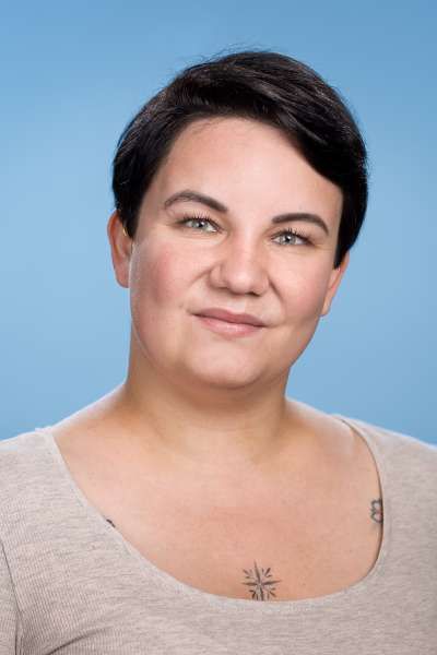 Eine Frau mit kurzen Haaren und einer Tätowierung auf der Brust lächelt in die Kamera.
