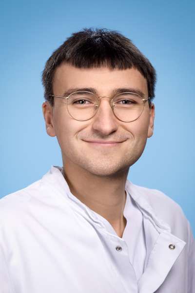 Ein junger Mann mit Brille und weißem Laborkittel lächelt in die Kamera.