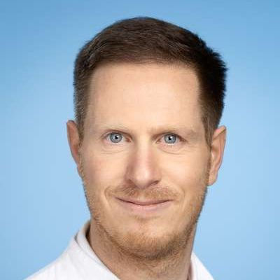 Ein Mann mit Bart und einem weißen Hemd lächelt in die Kamera.