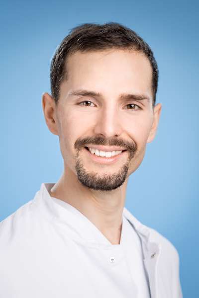 Ein Mann mit Bart trägt einen weißen Laborkittel und lächelt in die Kamera.