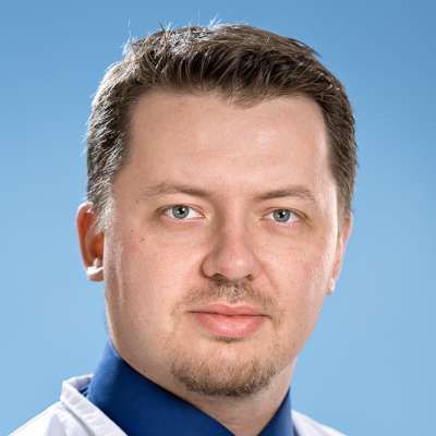 Ein Mann mit Bart trägt einen weißen Mantel und ein blaues Hemd.