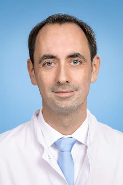 Ein Mann in einem weißen Kittel und einer blauen Krawatte lächelt in die Kamera.