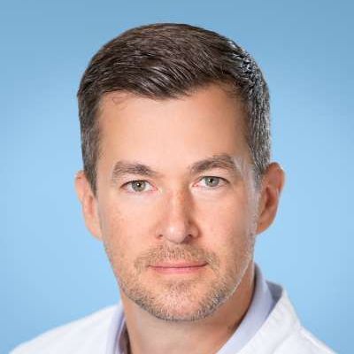 Ein Mann mit Bart trägt einen weißen Kittel und lächelt in die Kamera.