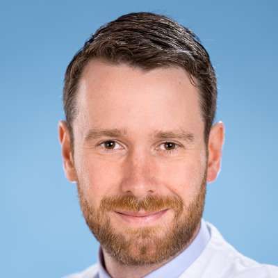 Ein Mann mit Bart trägt einen weißen Kittel und lächelt in die Kamera.