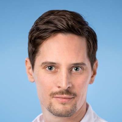 Ein Mann mit Bart schaut vor einem blauen Hintergrund in die Kamera.