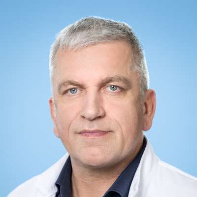 Ein Mann in einem weißen Kittel und einem blauen Hemd schaut in die Kamera.