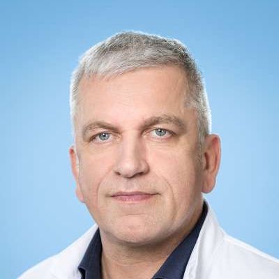 Ein Mann in einem weißen Kittel und einem blauen Hemd schaut in die Kamera.