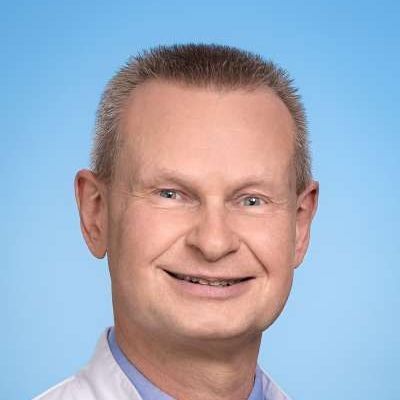 Ein Mann in einem weißen Kittel und einem blauen Hemd lächelt in die Kamera.