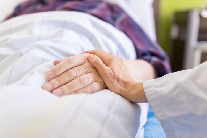 Die Hand einer medizinischen Fachkraft hält sanft die Hand eines Patienten, der in einem Krankenhausbett ruht.