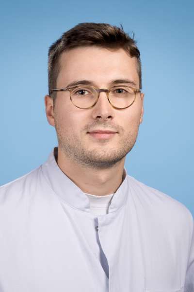 Ein Mann mit Brille und weißem Kittel steht vor einem blauen Hintergrund.