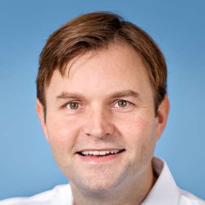 Ein Mann mit braunem Haar und weißem Hemd lächelt in die Kamera.