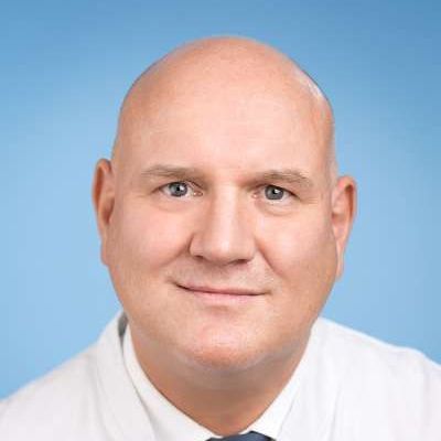 Ein kahlköpfiger Mann in weißem Kittel und Krawatte lächelt in die Kamera.