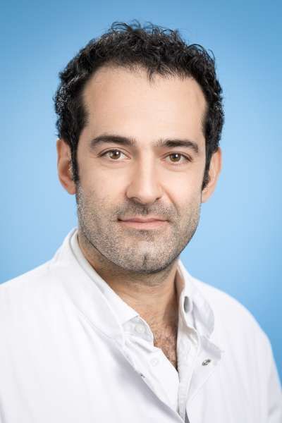 Ein Mann mit Bart trägt einen weißen Kittel und lächelt in die Kamera.