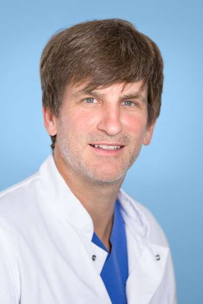 Ein Mann in einem weißen Kittel und einem blauen OP-Kittel lächelt in die Kamera