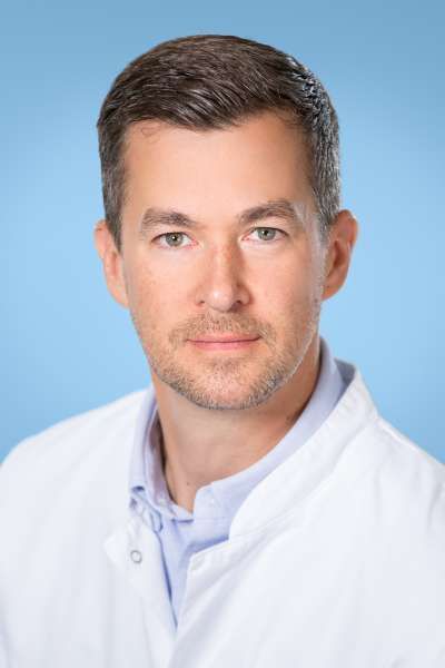 Ein Mann im weißen Kittel steht vor einem blauen Hintergrund.