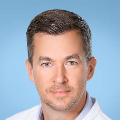 Ein Mann mit Bart trägt einen weißen Kittel und lächelt in die Kamera.