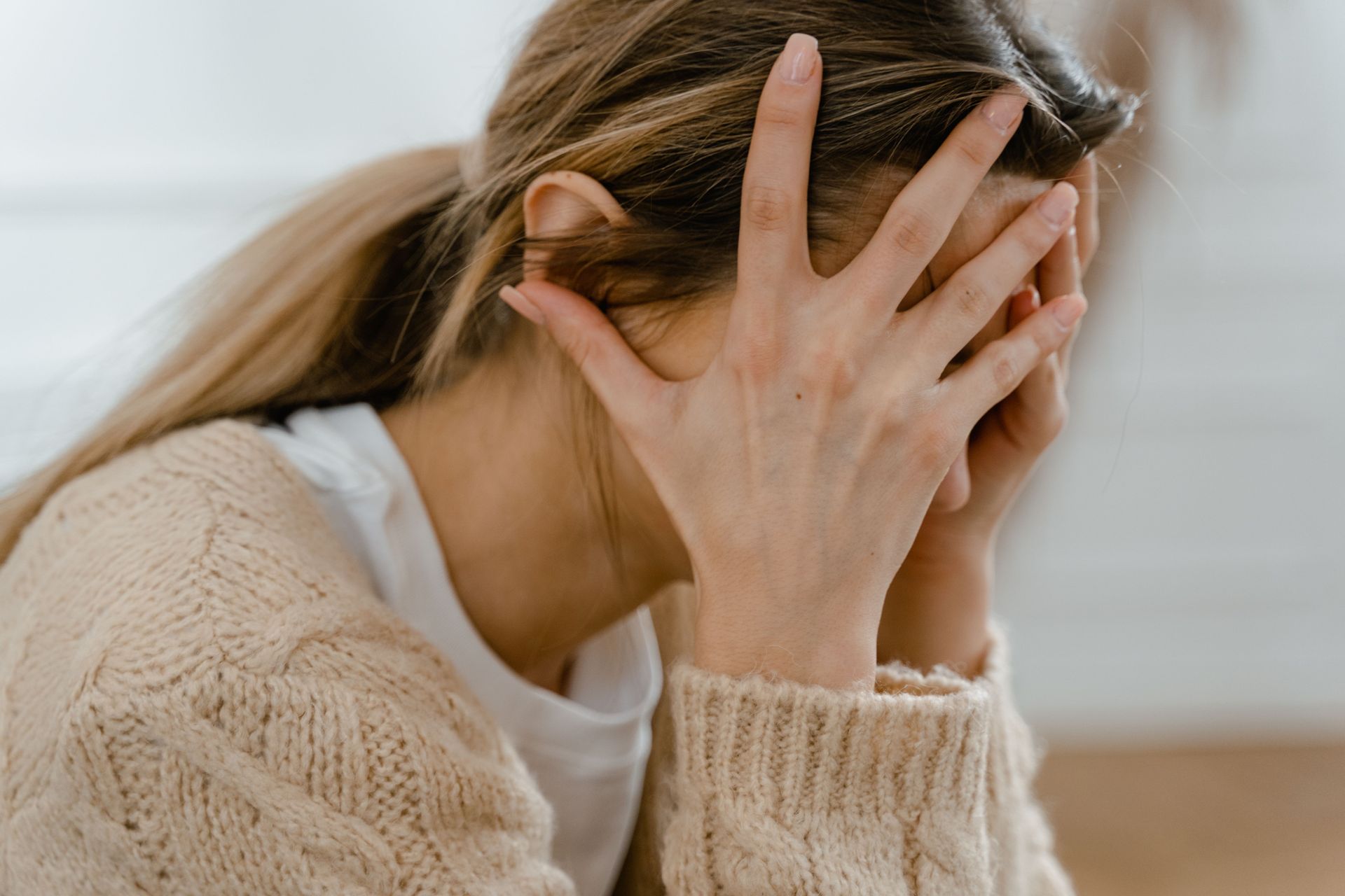 Person mit Händen, die das Gesicht bedecken, in einem beigen Pullover, sitzt in einem Innenraum in einer verzweifelten Pose