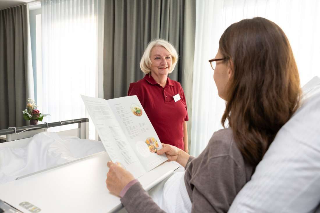 Eine Frau sitzt in einem Krankenhausbett und spricht mit einer Krankenschwester.