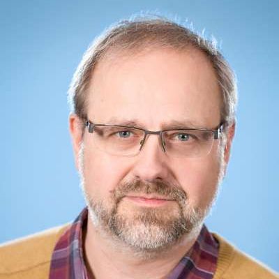 Ein Mann mit Bart und Brille trägt ein kariertes Hemd und einen gelben Pullover.