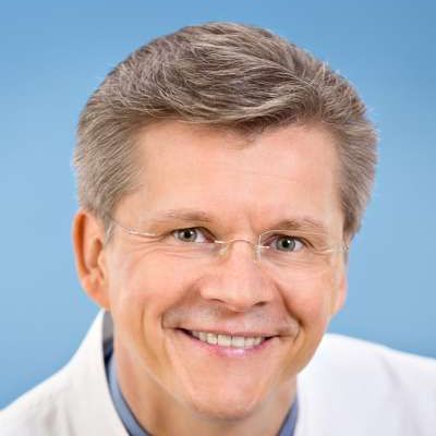 Ein Mann im weißen Kittel und mit Brille lächelt in die Kamera.