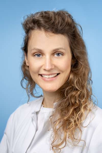 Eine Frau mit lockigem Haar trägt einen weißen Laborkittel und lächelt.