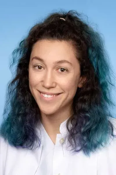 Eine Frau mit blauen Haaren und weißem Hemd lächelt in die Kamera.