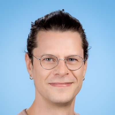 Ein Mann mit Brille und Ohrringen lächelt in die Kamera.