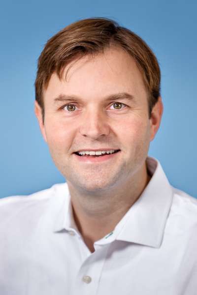 Ein Mann in einem weißen Hemd lächelt vor einem blauen Hintergrund in die Kamera.