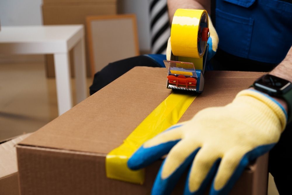 Person taping a cardboard box with a yellow tape dispenser, wearing gloves.