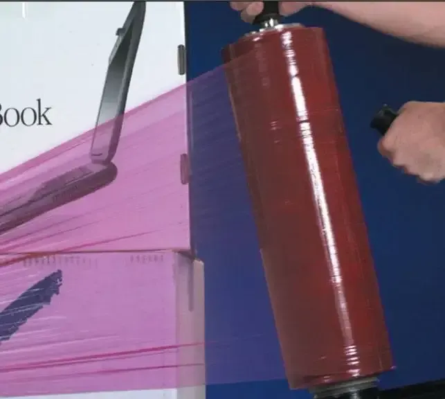 Person wrapping a box with pink plastic using a handheld dispenser, indoors.