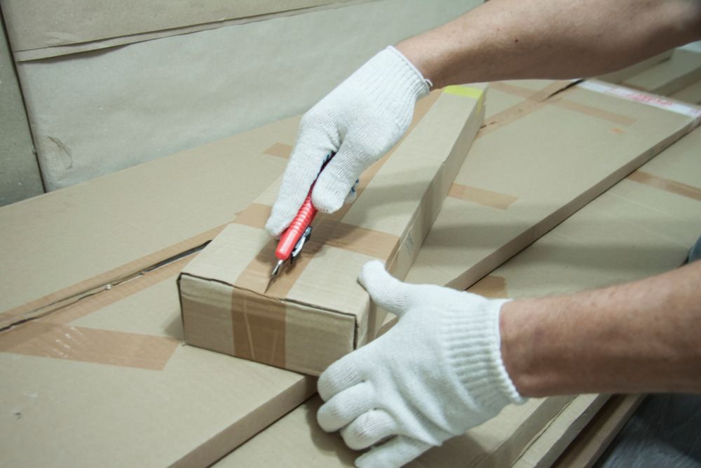 Person wearing gloves cutting a small cardboard box with a utility knife, surrounded by other boxes.