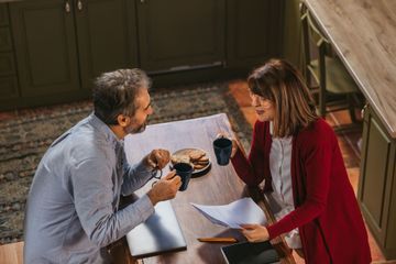 A couple sits at a table, drinking coffee and looking at papers. Kitchen setting.