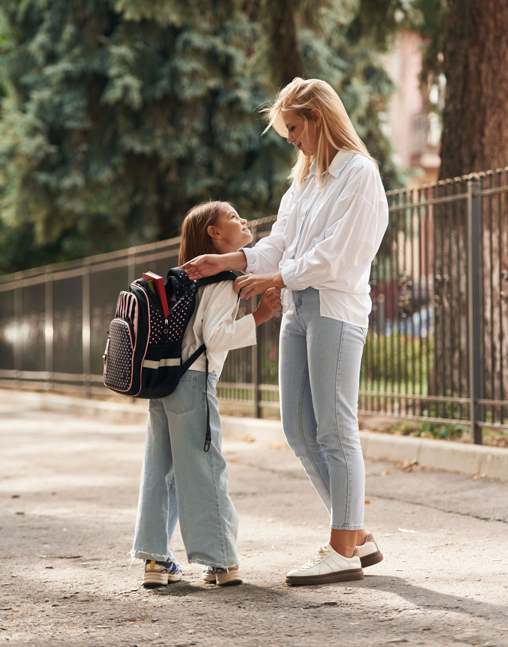 Woman adjusting backpack for child near a fence, both wearing jeans and white tops, on a paved path.
