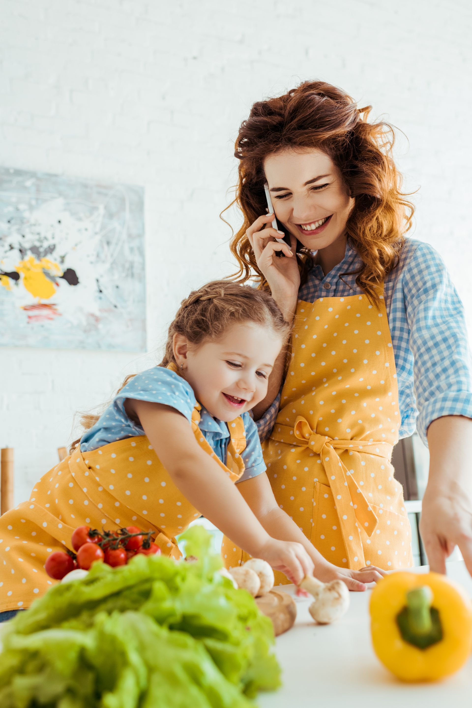 Woman on phone smiles while daughter reaches for mushrooms; both wearing yellow aprons in kitchen.