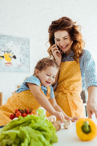 Woman on phone smiles while daughter reaches for mushrooms; both wearing yellow aprons in kitchen.