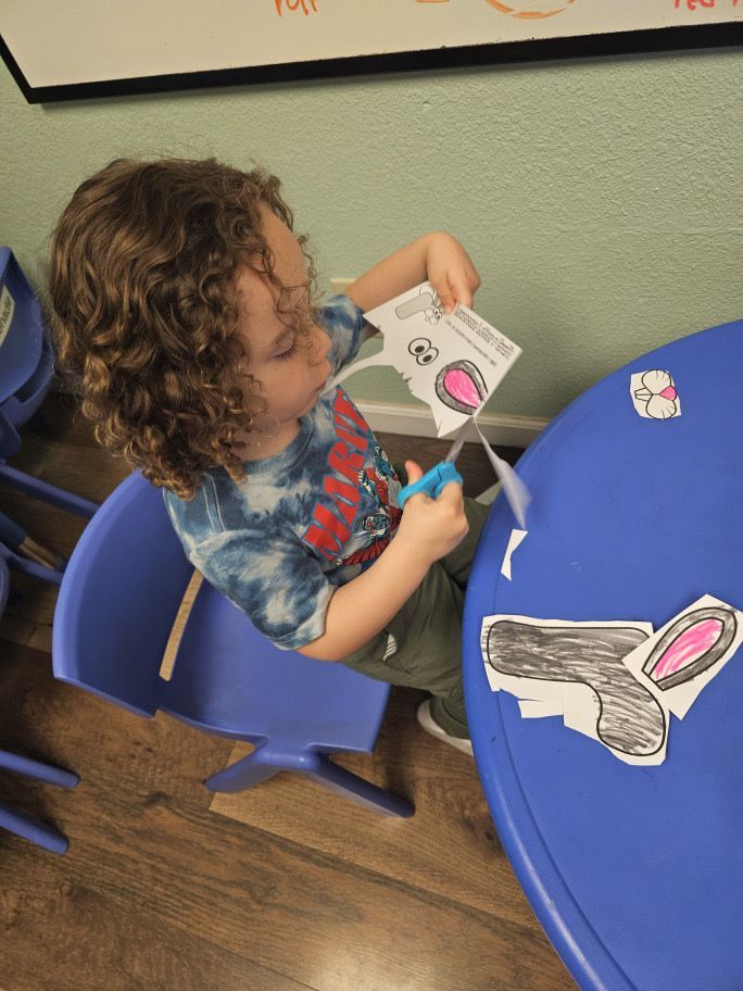A young boy in a tie dye shirt is cutting a piece of paper with scissors