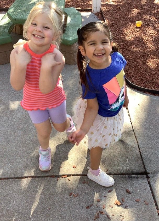Two little girls are standing next to each other on a sidewalk and smiling.