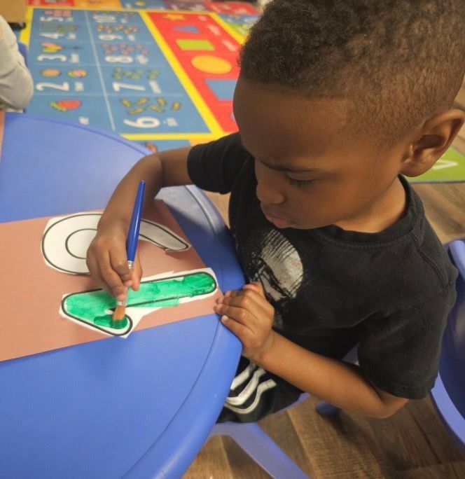A young boy sits at a table drawing a letter g on a piece of paper