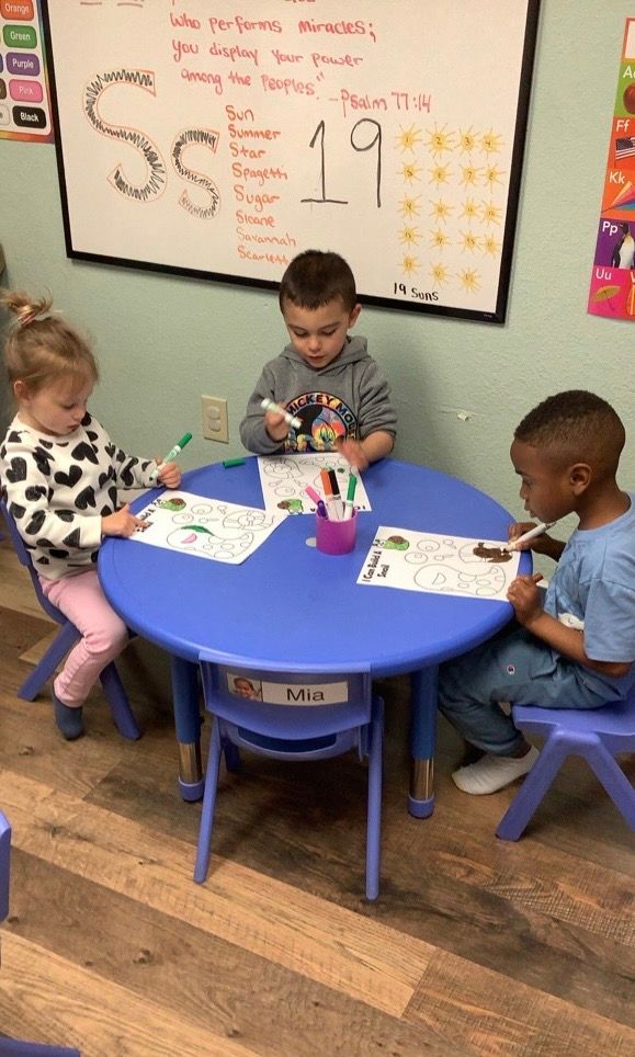 Three children are sitting at a blue table in front of a whiteboard.