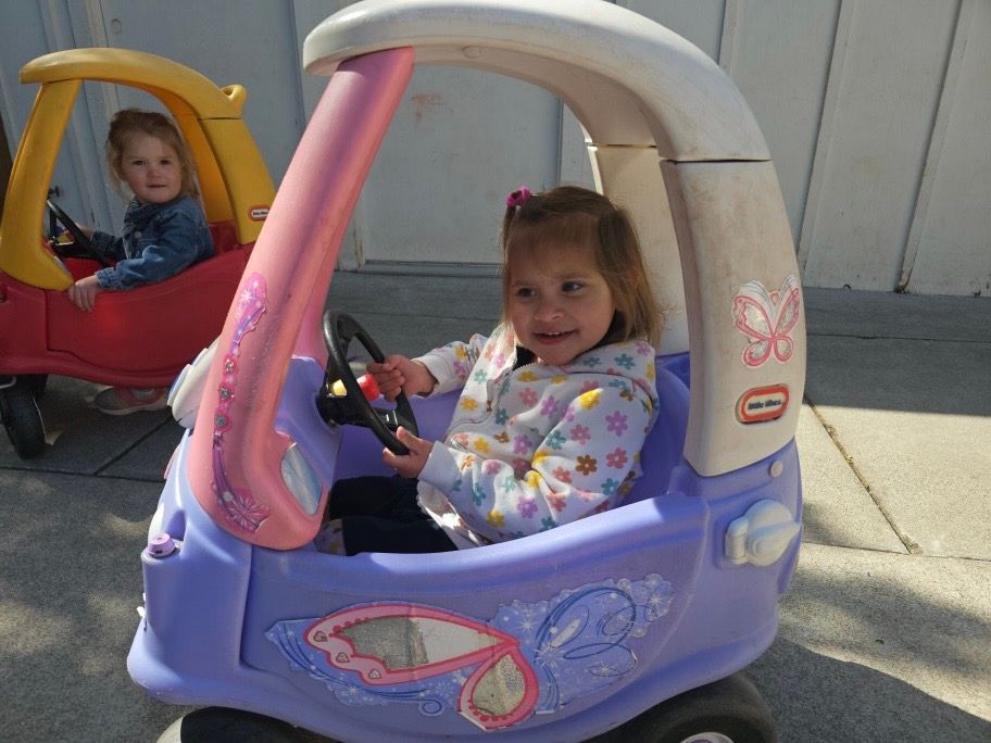 A little girl is sitting in a cozy coupe toy car