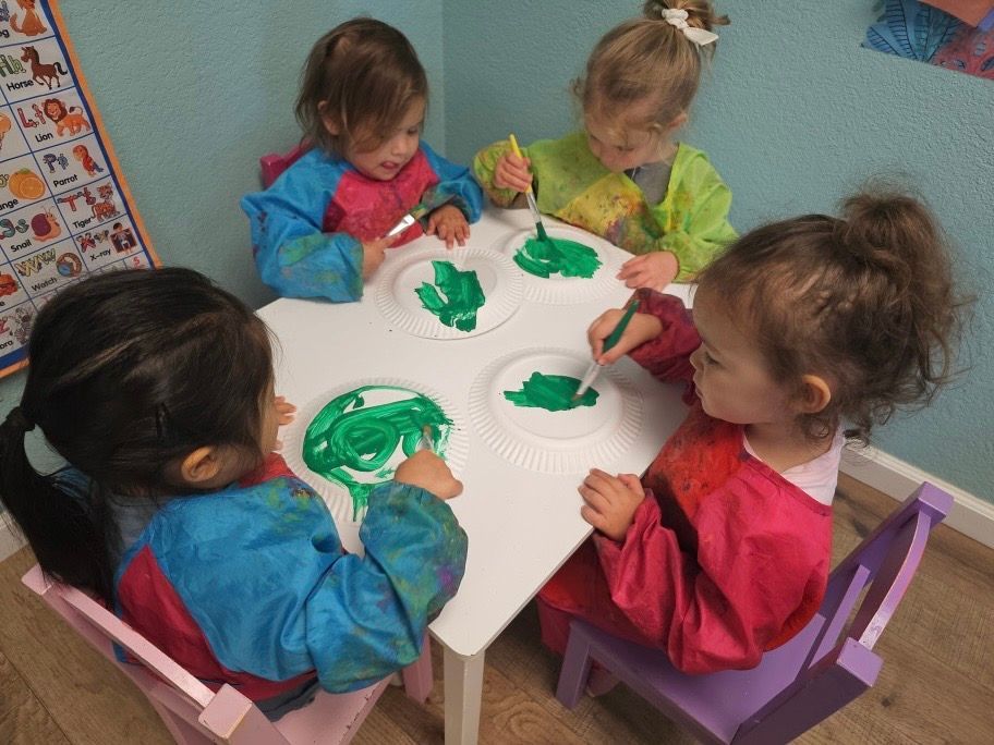 Four little girls are sitting at a table painting with green paint