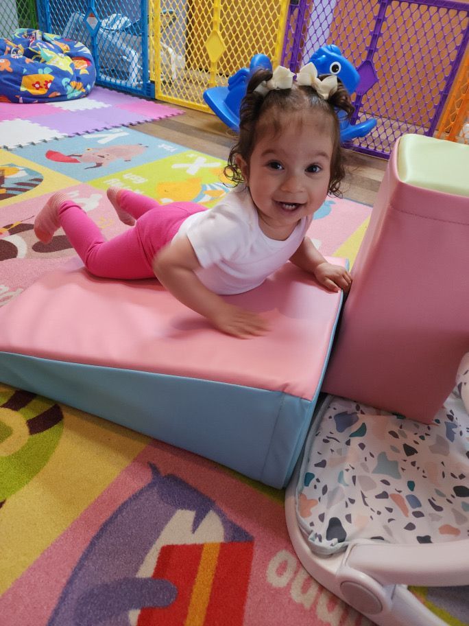A little girl is crawling on a pink cushion on the floor.