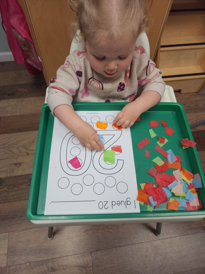 A little girl is sitting at a table playing with a piece of paper with the number 6 on it