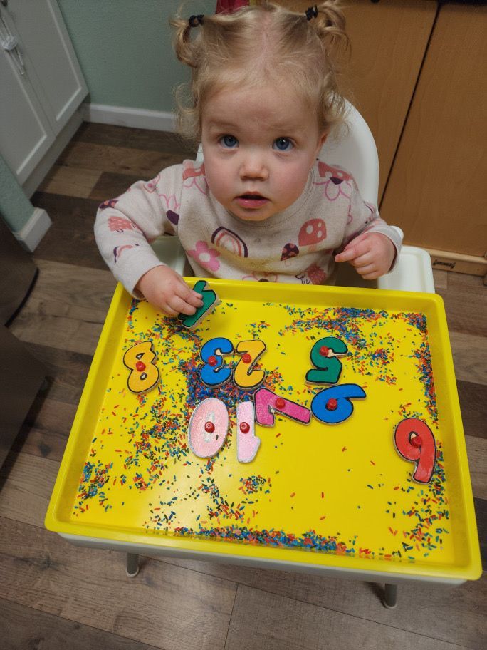 A little girl is sitting in a high chair playing with sprinkles and numbers.