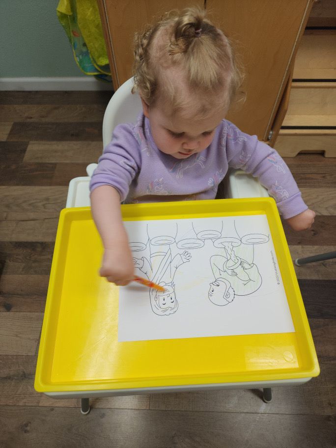 A little girl is sitting in a high chair drawing on a yellow tray
