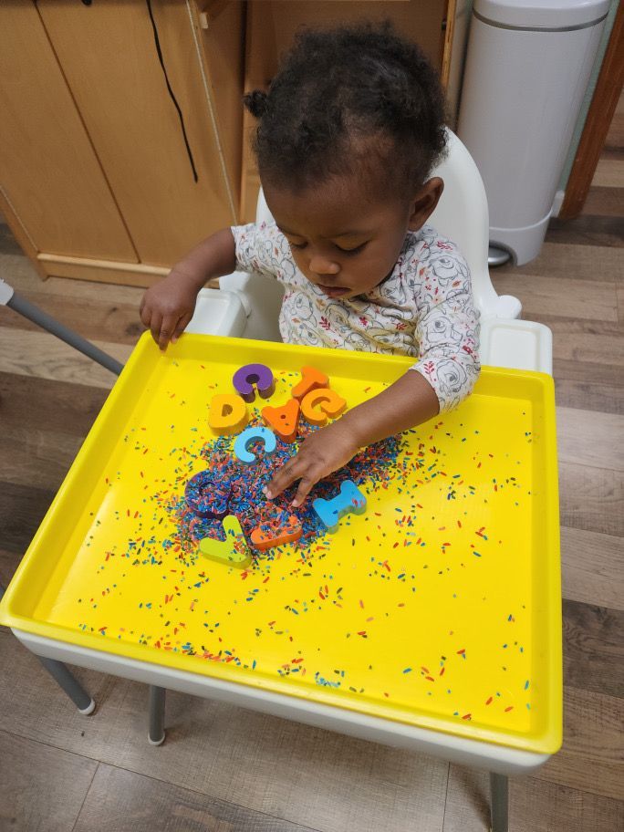 A baby is sitting in a high chair playing with sprinkles and letters.