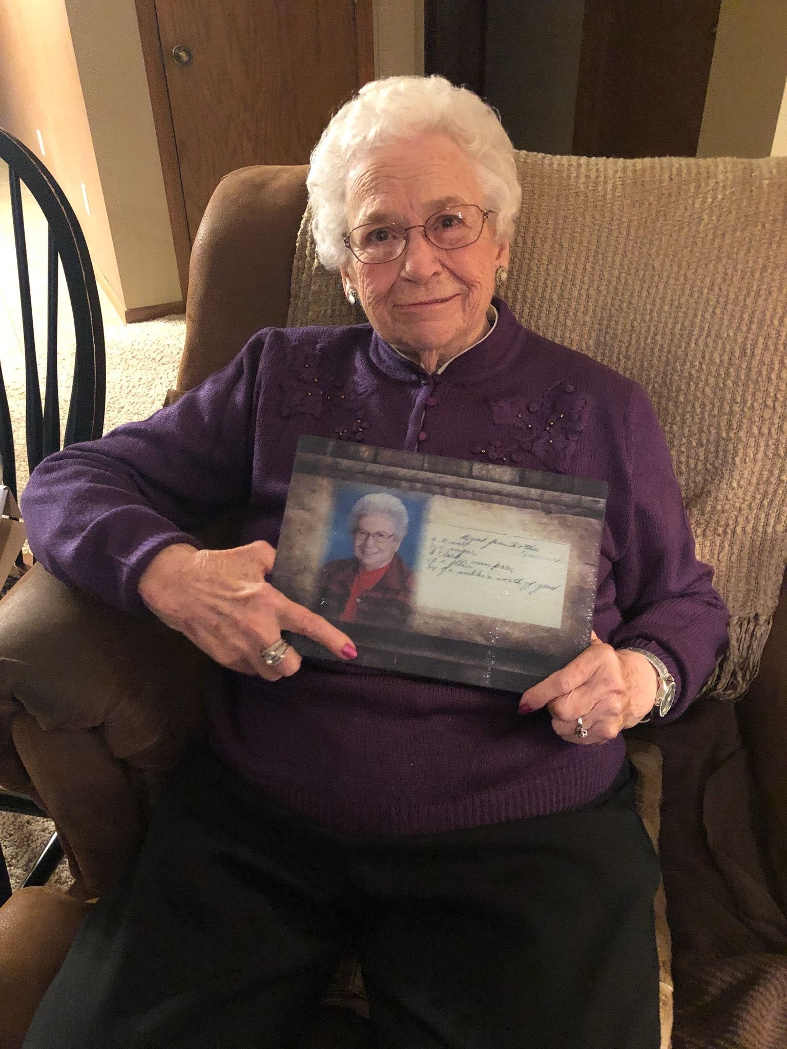 Older woman in purple sweater holds photo frame, seated in a chair, smiling.