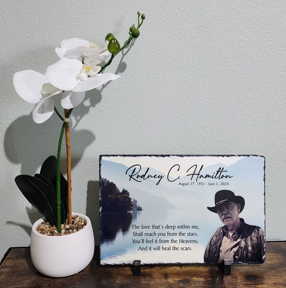 Memorial plaque with photo of Rodney C. Hamilton, next to white orchid plant in pot.